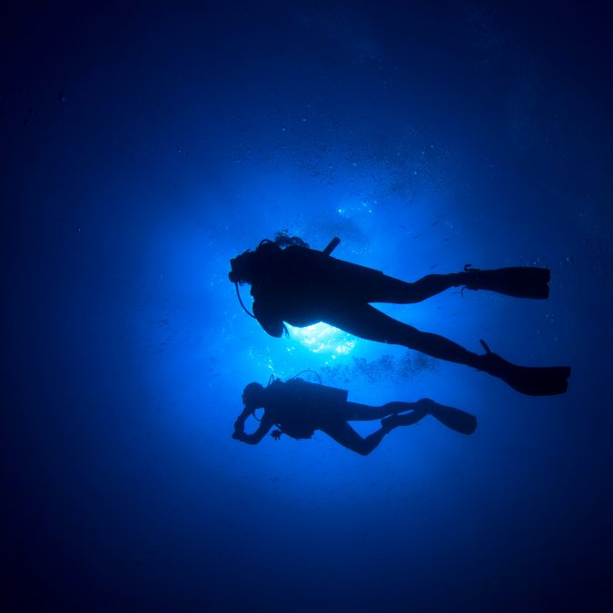 2 scuba divers in the deep blue with sun behind them 
