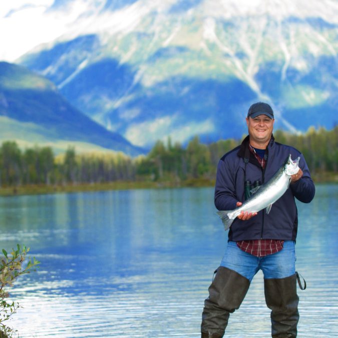 fisherman in alaska holding a salmon with a lake behind him